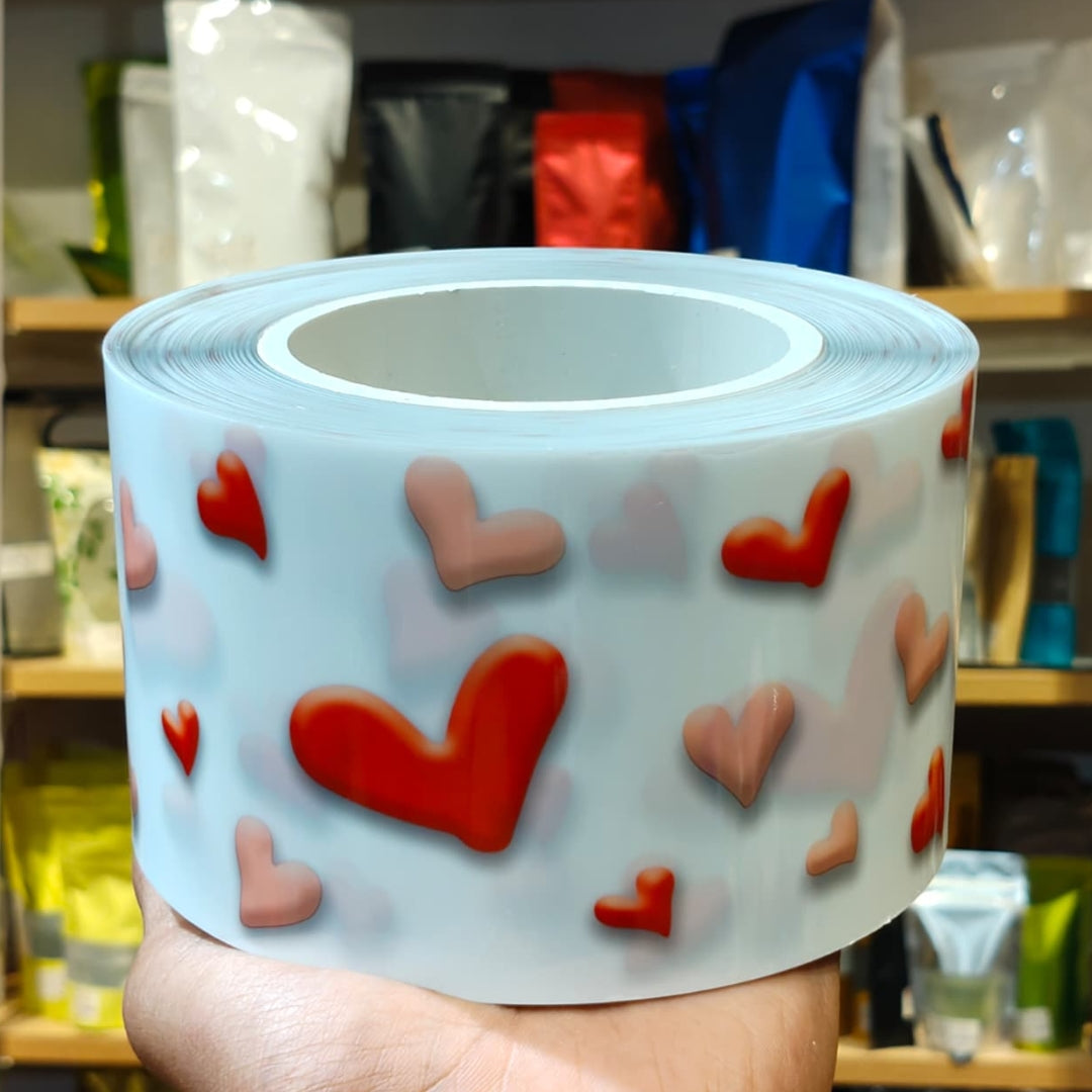 A roll of clear tape with a pattern of red and pink hearts, held in a hand against a blurred background of shelves with bags and boxes.