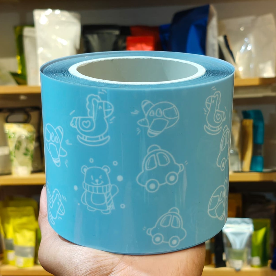 A hand holds a roll of light blue tape printed with white cartoon-style outlines of animals, vehicles, and objects, with a blurred store shelf in the background.
