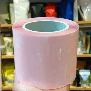 A hand holds a large roll of light pink tape with a white repeating pattern of strawberries, bows, and stars. The background shows a blurred shop shelf with various colorful items.