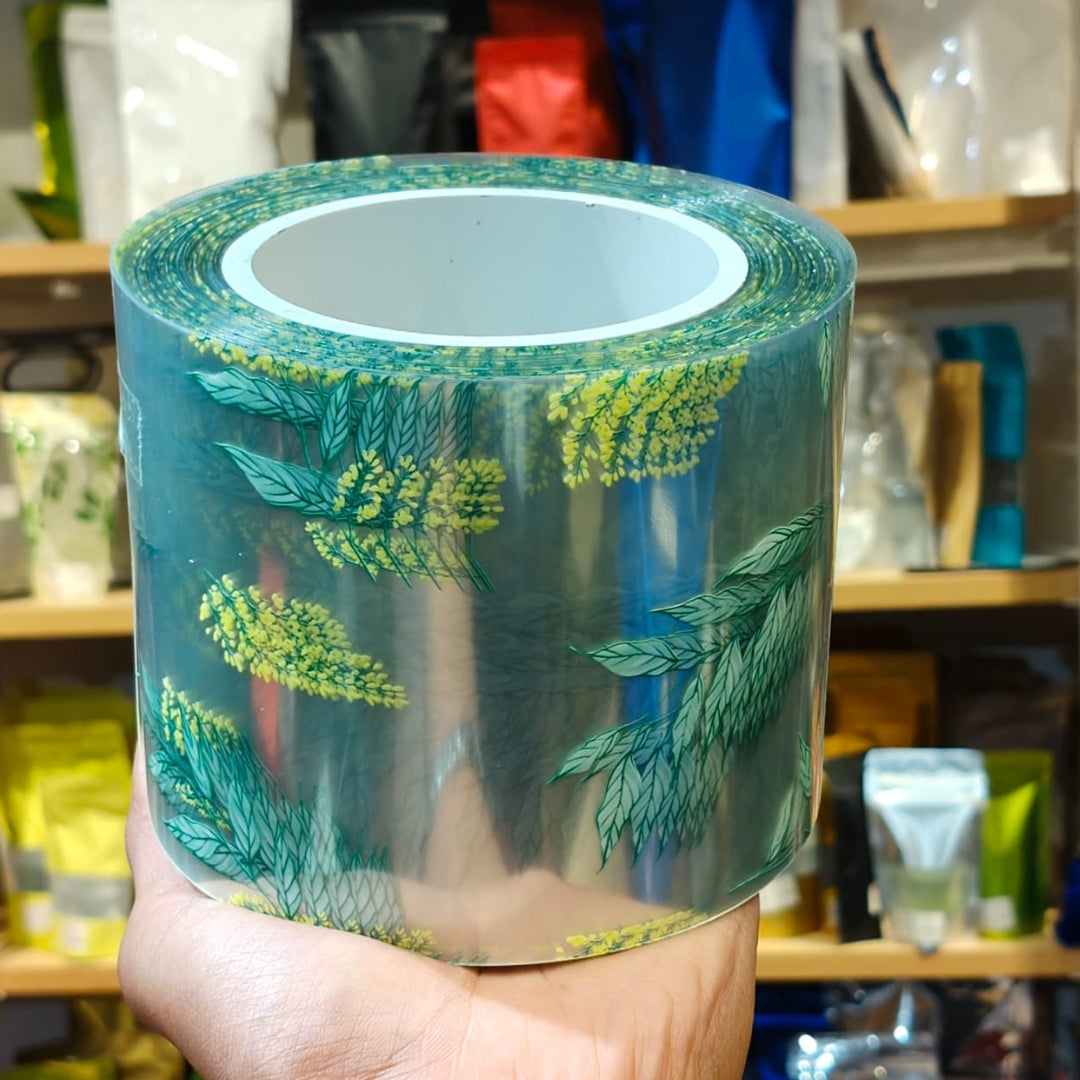 A hand holds a roll of clear adhesive tape featuring a printed pattern of green leaves and yellow flowers, with a white inner core, set against a blurred background of shelves containing various items.