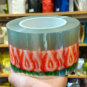 A hand holding a roll of clear adhesive tape printed with a repeating strawberry slice pattern in red, pink, and green, with a white inner core.