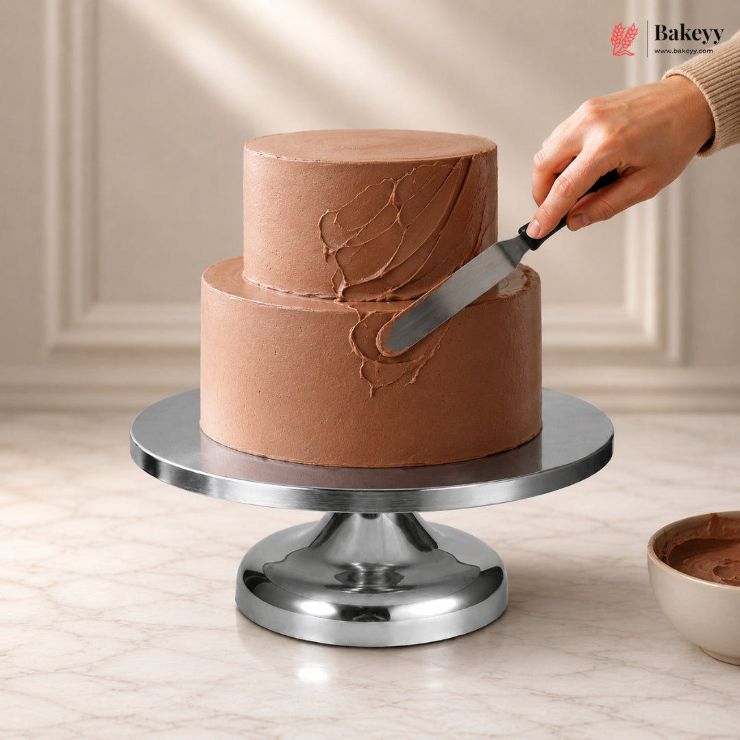 Chocolate cake being frosted on a metal cake stand with a hand holding a spatula.