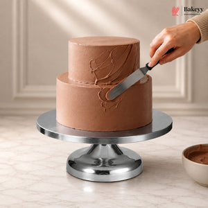 Chocolate cake being frosted on a metal cake stand with a hand holding a spatula.