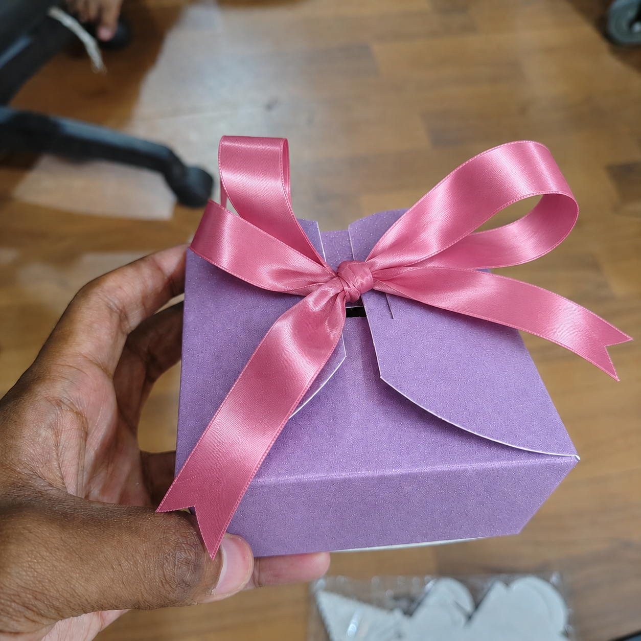 Purple gift box with a pink ribbon held by a hand on a wooden floor.
