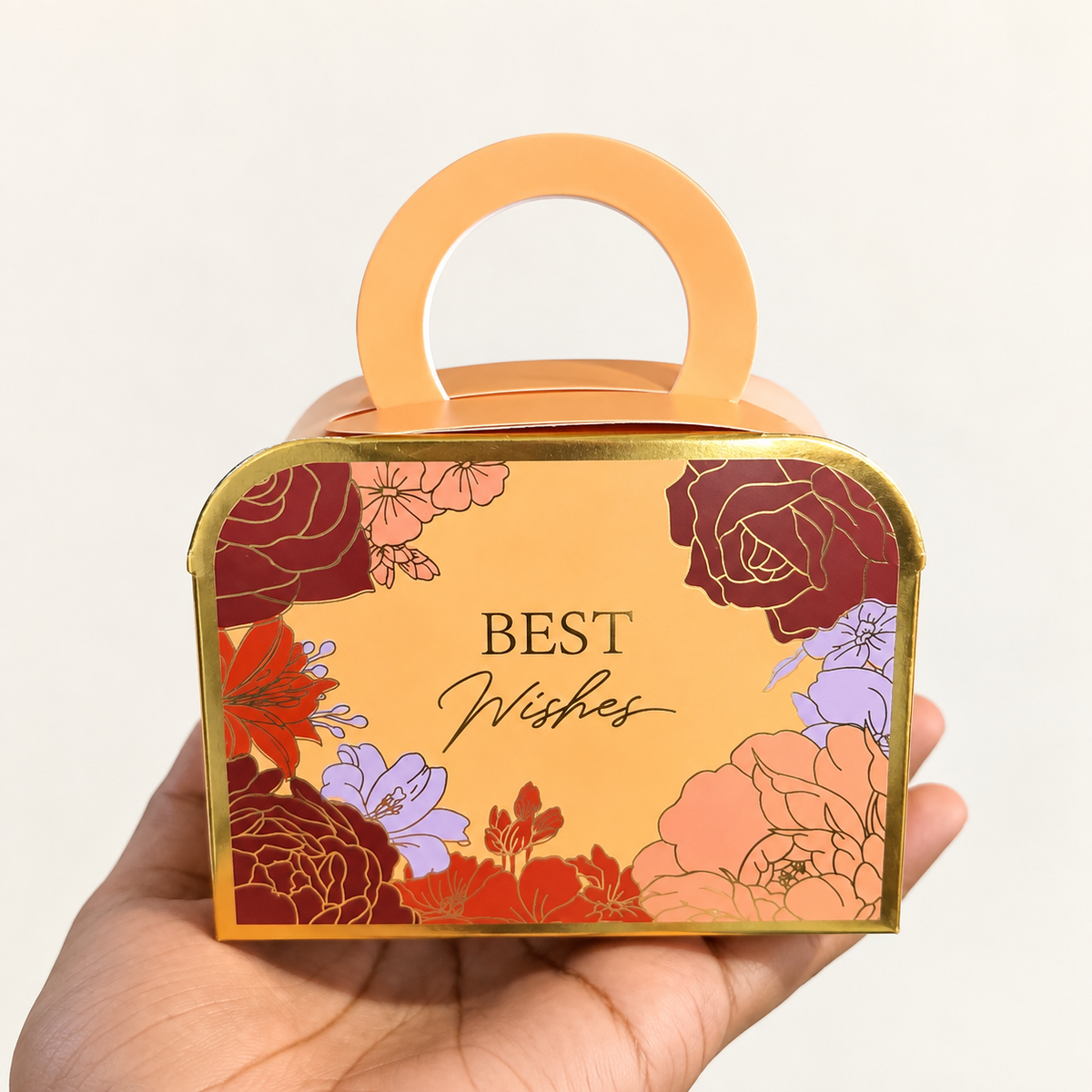 A hand holding a decorative gift box with a handle. The box has a warm peach background with gold foil borders and colorful floral illustrations in red, orange, purple, and pink.