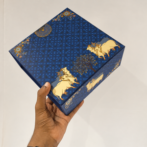 A hand holding a square blue decorative box with gold illustrations of animals and floral motifs against a plain light background.