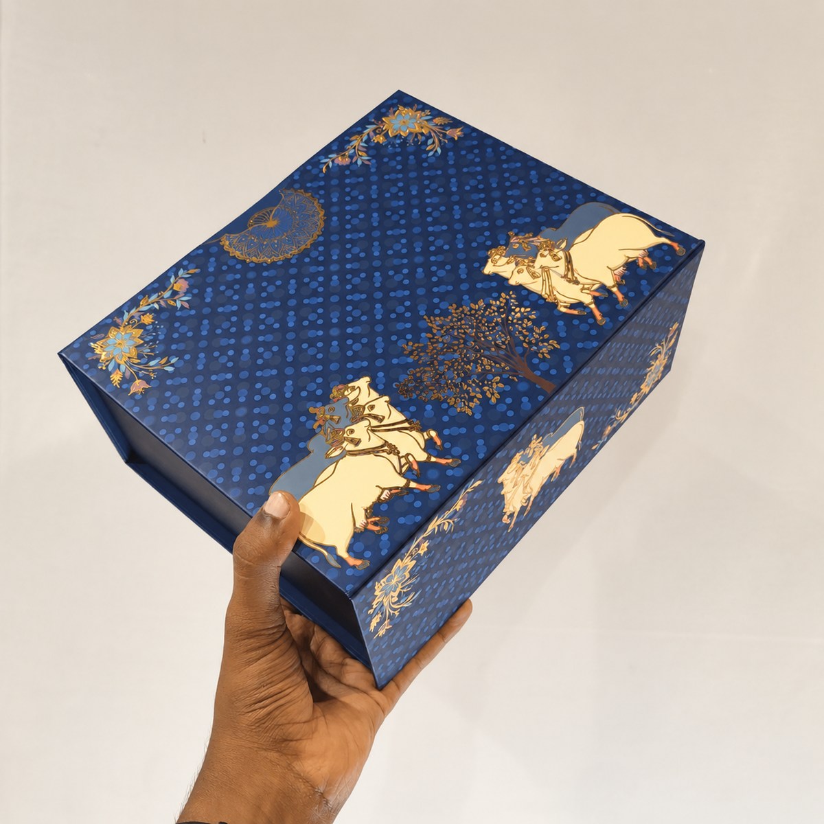 A hand holding a rectangular blue decorative box with gold and cream illustrations of animals, trees, and floral motifs, photographed against a plain light background.