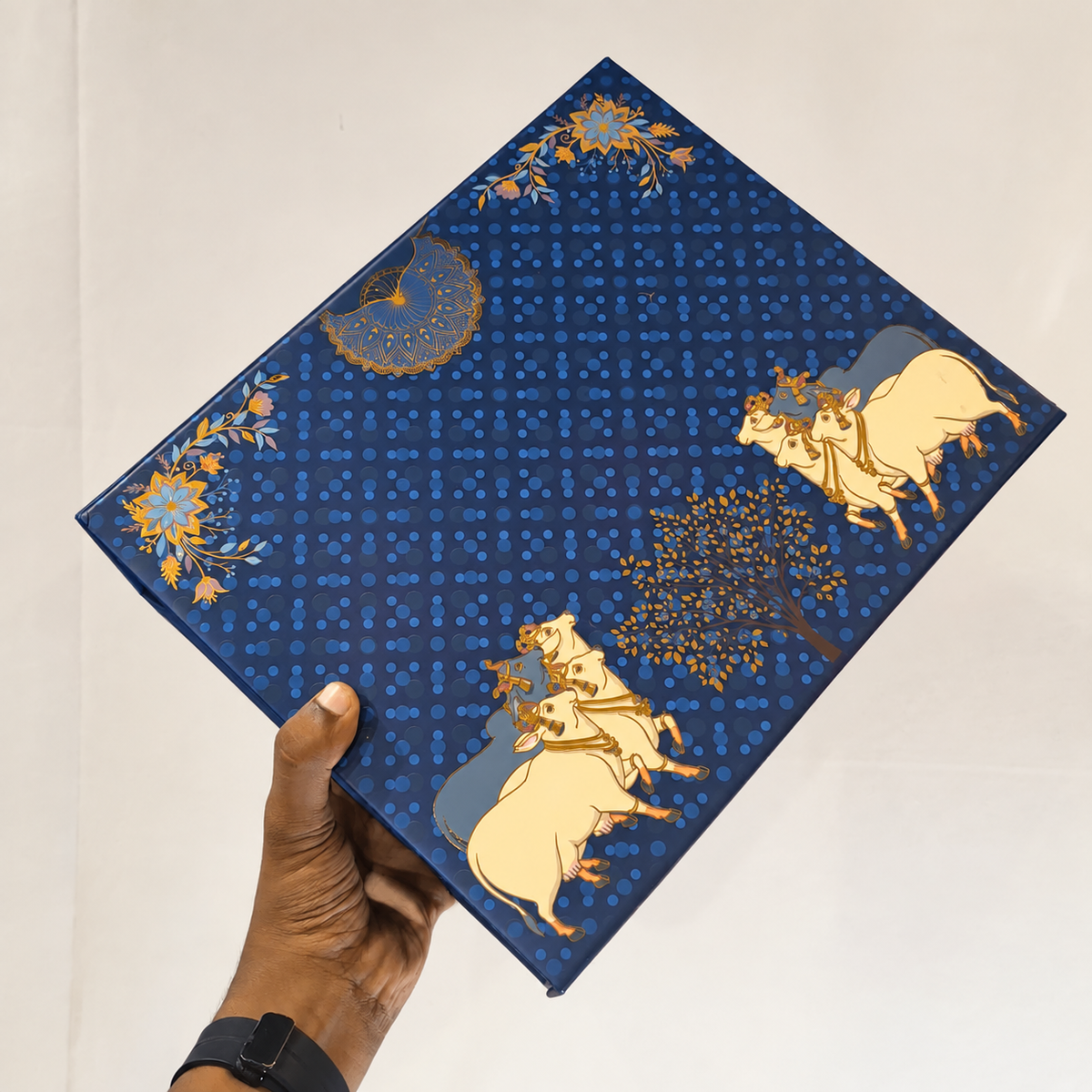 A hand holds a square fabric item, likely a pocket square or small scarf, featuring a deep blue background with a repeating small pattern, gold floral accents, and cream and blue illustrations of horses and a tree.