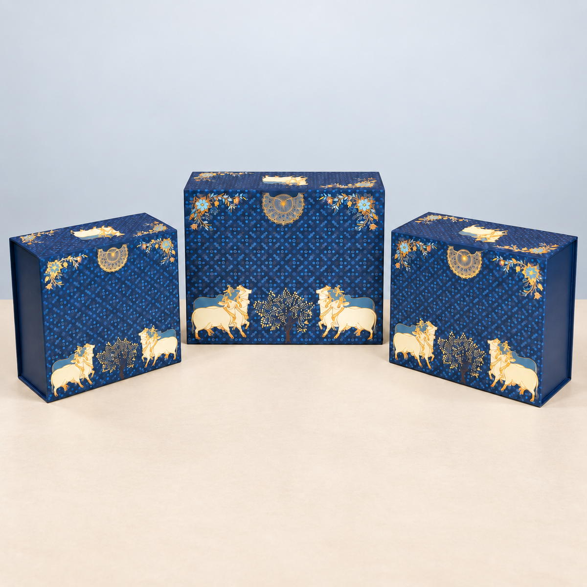 Three decorative rectangular boxes in different sizes, arranged on a light beige surface against a pale blue background. The boxes are dark blue with an allover geometric pattern and feature cream and gold illustrations of lions, trees, and circular motifs.