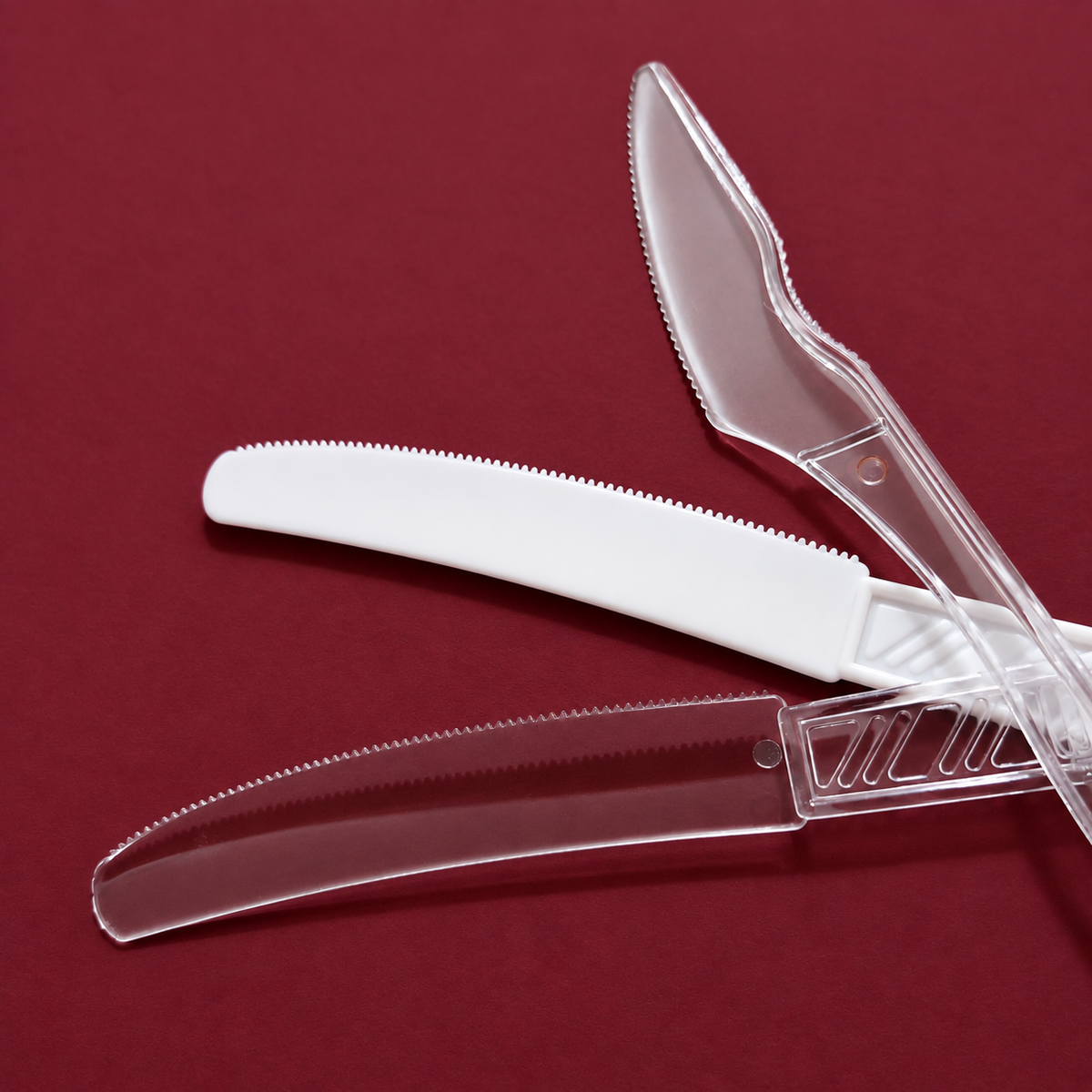 Two plastic plastic-knife style utensils with serrated edges, one white and one transparent, arranged on a dark red background.