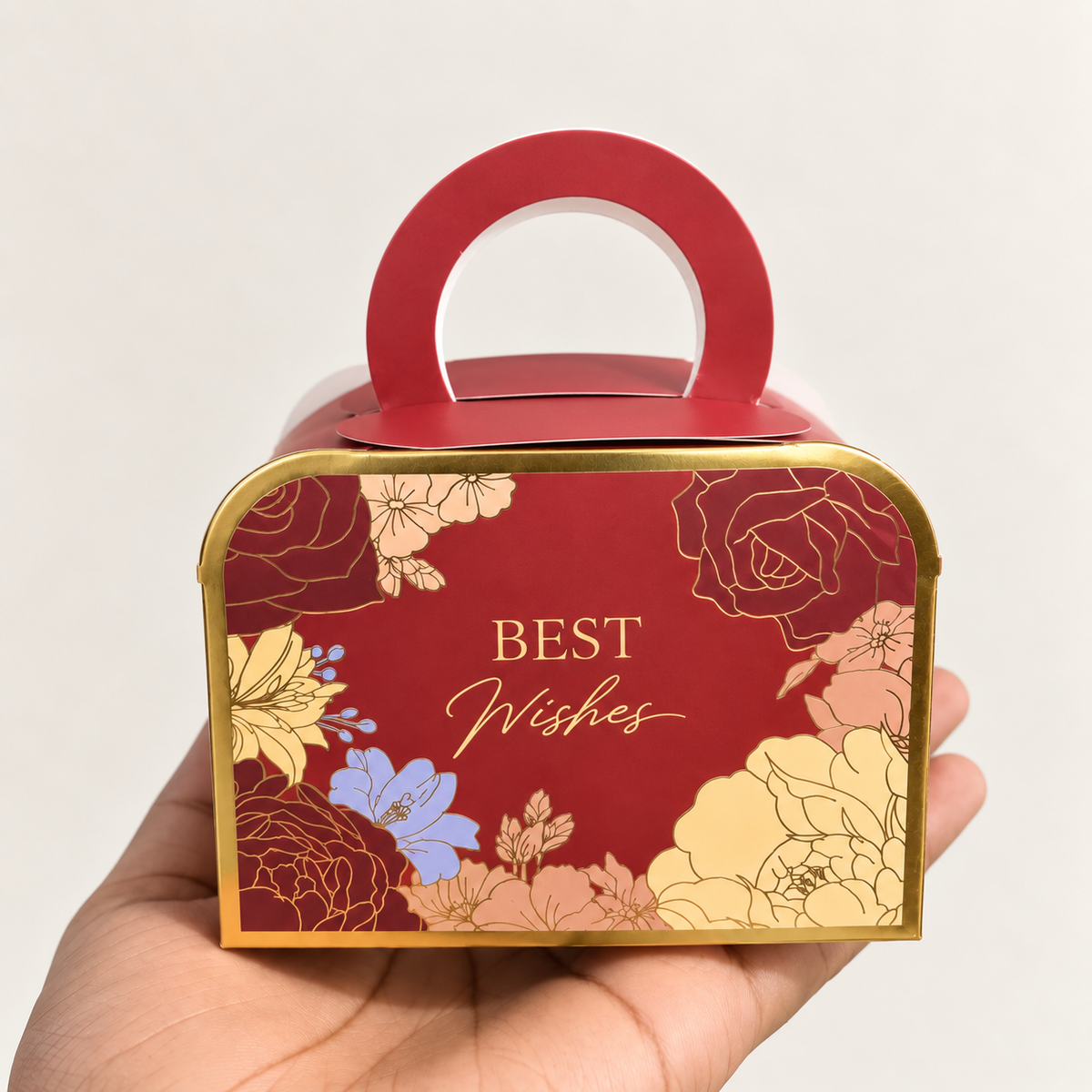 A hand holding a red and gold gift box with a handle, decorated with floral illustrations and the words 'BEST Wishes' in gold lettering.