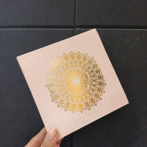 A hand holding a square pale pink box or card featuring a large metallic gold mandala design on the front, set against a dark gray tiled background.