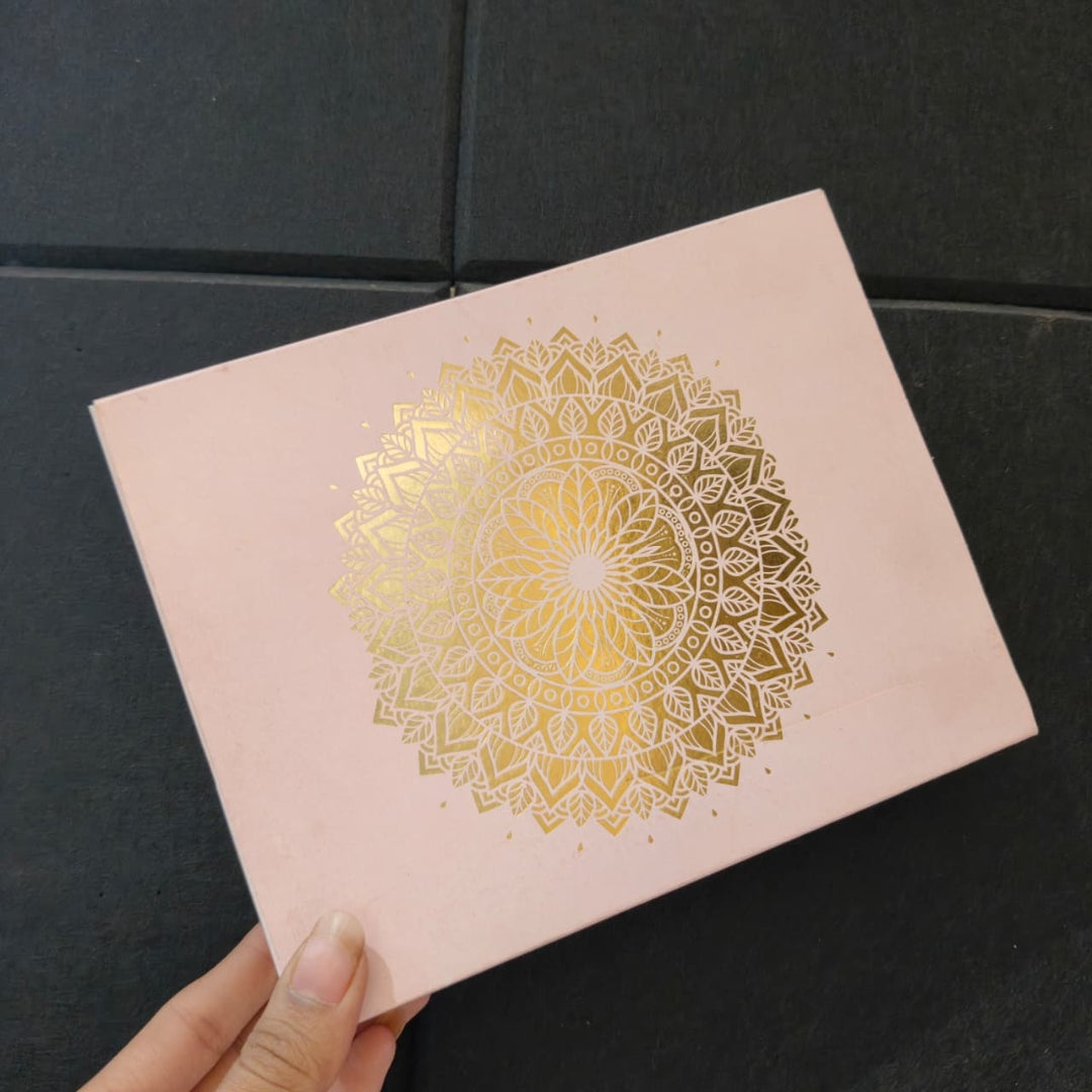 A hand holding a square pink notebook or journal with a large metallic gold mandala design on the cover, set against a dark textured background.