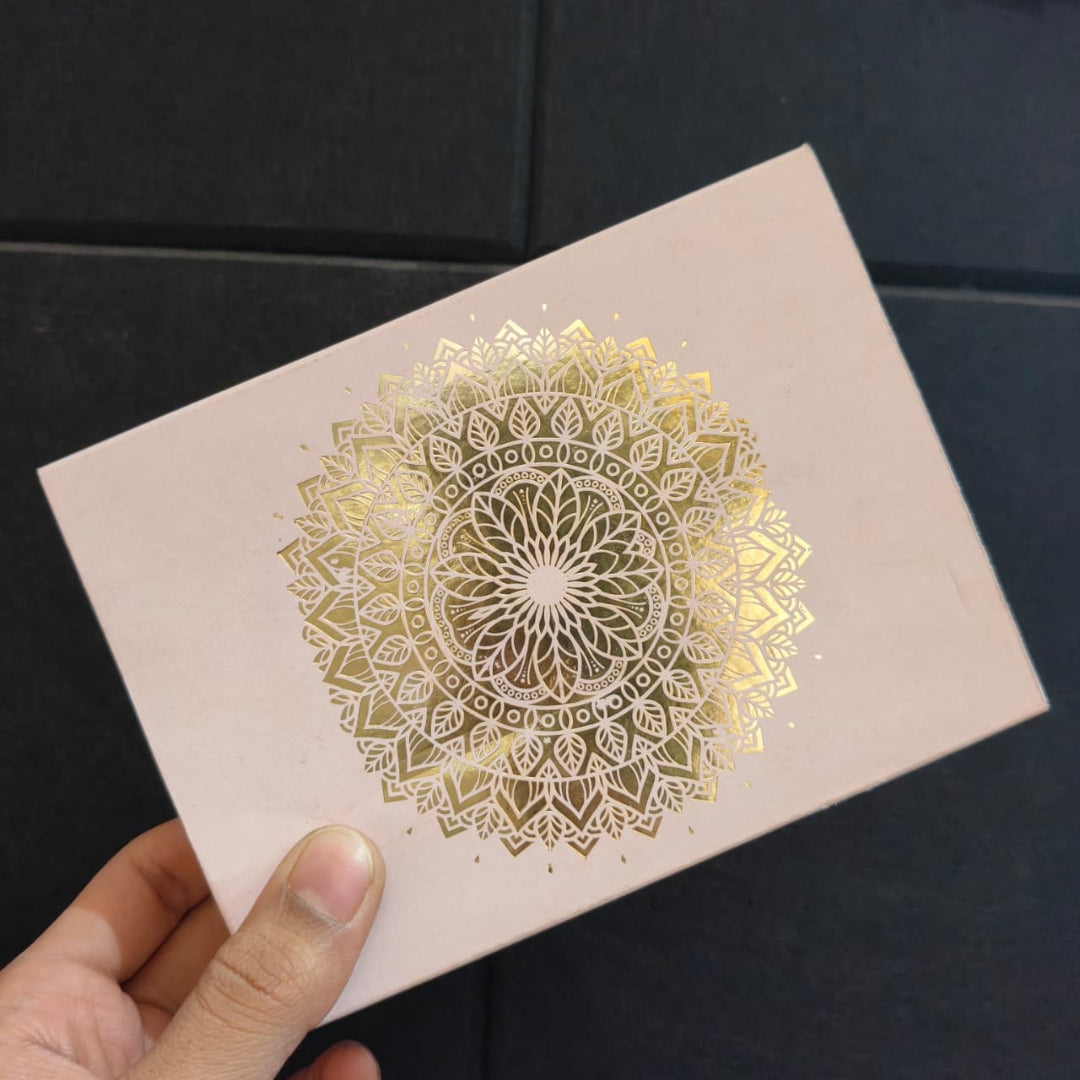 A hand holding a square blush pink card or notebook cover featuring a large metallic gold mandala design in the center, set against a dark tiled background.