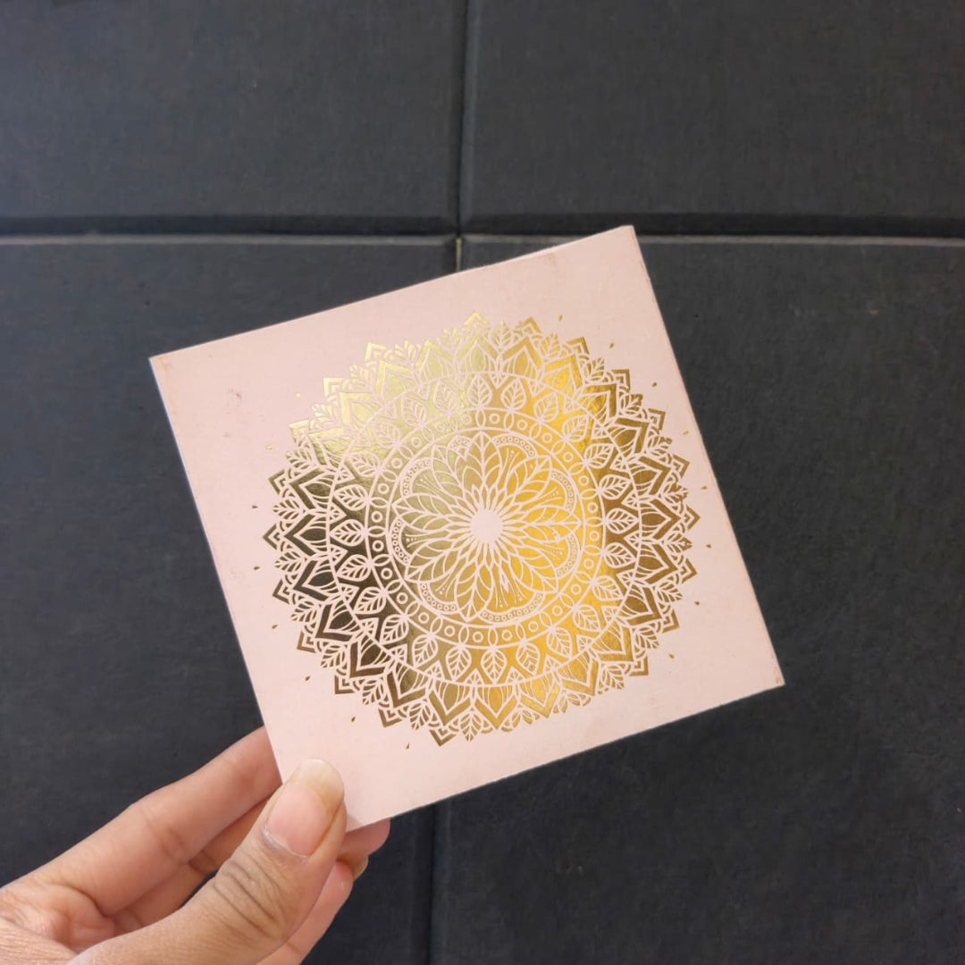 A hand holding a square pink card featuring a metallic gold mandala design, set against a dark gray tiled background.