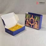 A blue and gold bakery gift box shown open with a white interior and gold tray insert, alongside a matching blue gift bag with gold rope handles and decorative gold artwork. The photo is on a light neutral studio surface with a gray backdrop.