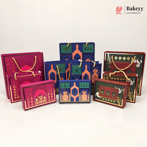 A collection of decorative gift boxes and shopping bags in rich jewel tones, including magenta, navy blue, green, and brown, featuring intricate architectural patterns and gold foil details. The products are arranged on a clean white studio background with a Bakeyy brand logo in the top right corner.