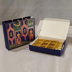 A navy blue gift box with gold foil compartments shown open next to a matching decorative gift bag with gold rope handles, on a light gray background.