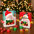 Two Christmas-themed candy boxes with Santa and snowman designs, filled with colorful candies, on a wooden surface with blurred Christmas lights in the background.