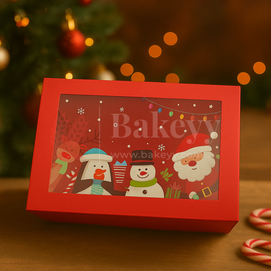 Red gift box with Christmas-themed design featuring Santa Claus, penguins, and a snowman, with 'bakeyy' branding, on a wooden surface with blurred Christmas tree and lights in the background.