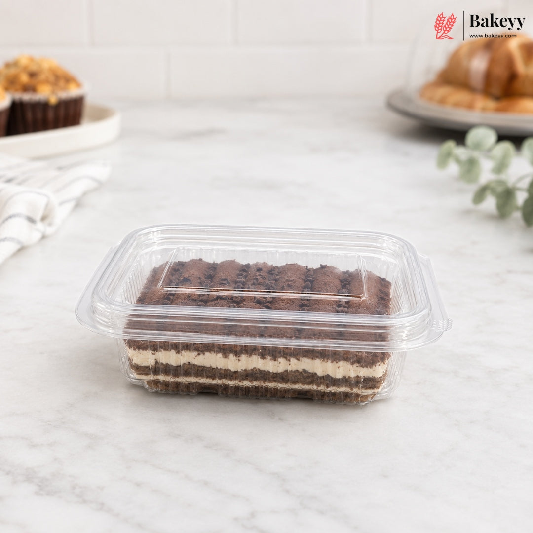 A rectangular clear plastic clamshell container holding a layered chocolate cake or pastry, placed on a light marble countertop with blurred bakery items in the background. The Bakeyy logo appears in the top right corner.
