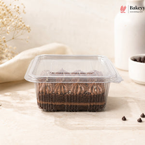 A clear plastic clamshell container holding a chocolate cake or dessert with piped chocolate frosting, set on a light beige surface with small decorative props and a Bakeey logo in the top right corner.