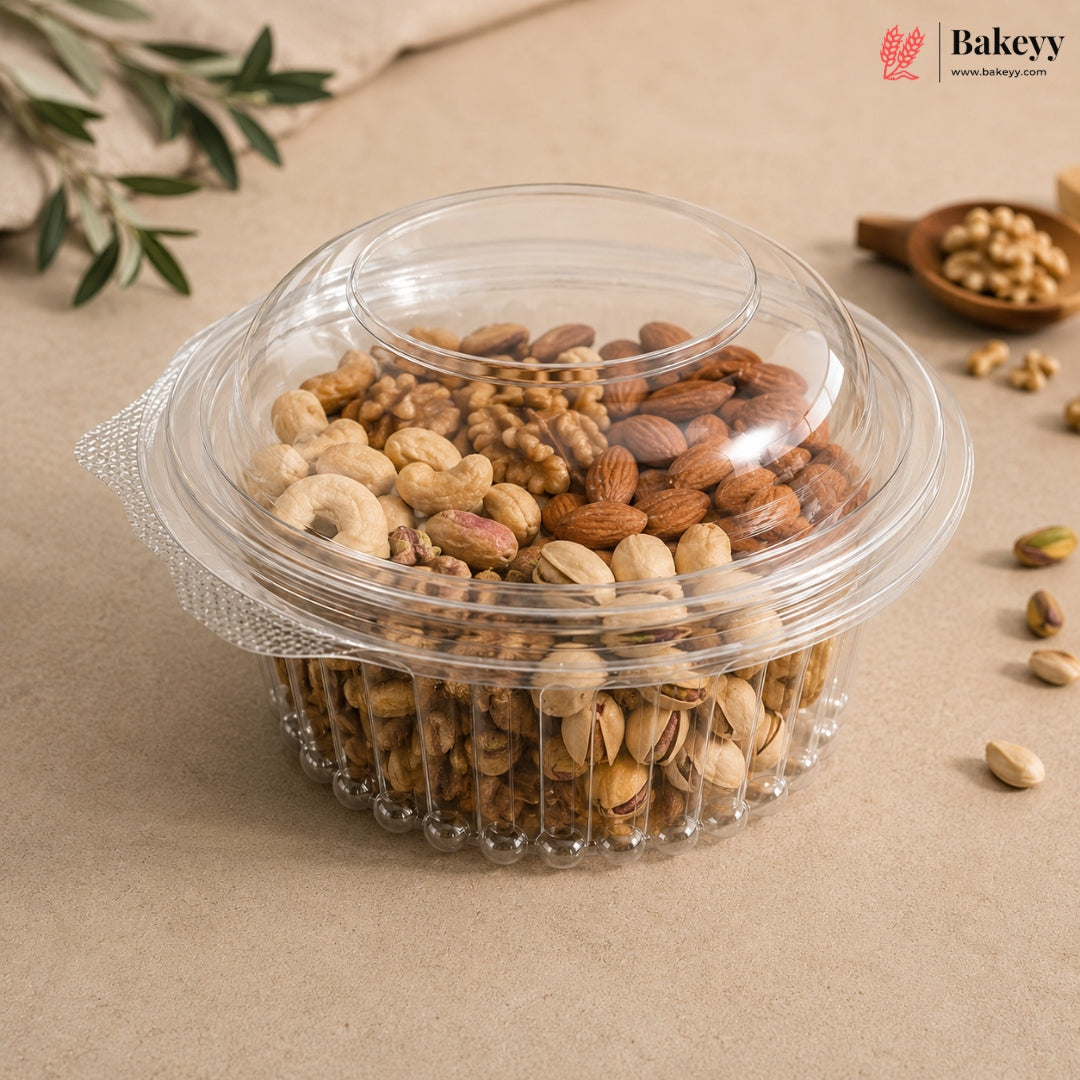 A clear plastic round container with a matching lid, filled with assorted nuts including almonds, cashews, and pistachios, photographed on a beige surface with small decorative props and a Bakeyy logo in the top right.
