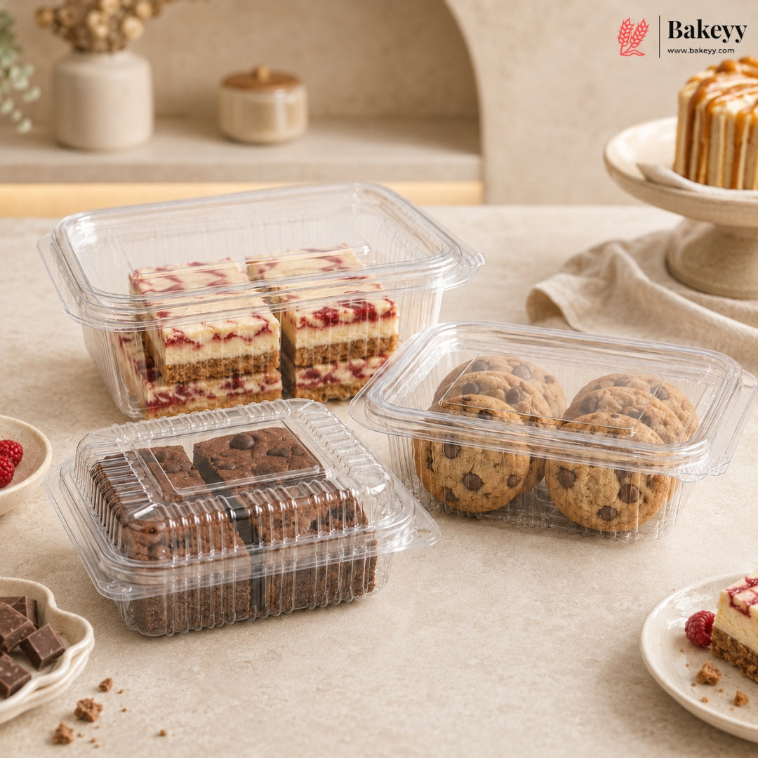 Three clear plastic food storage containers holding assorted desserts, including chocolate chip cookies, brownies, and layered cakes, arranged on a beige tabletop with a bakery logo in the top right corner.