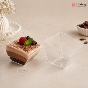 Two clear plastic dessert cups on a beige surface, with one cup filled with a layered chocolate dessert topped with a raspberry and mint, and the other cup shown empty.