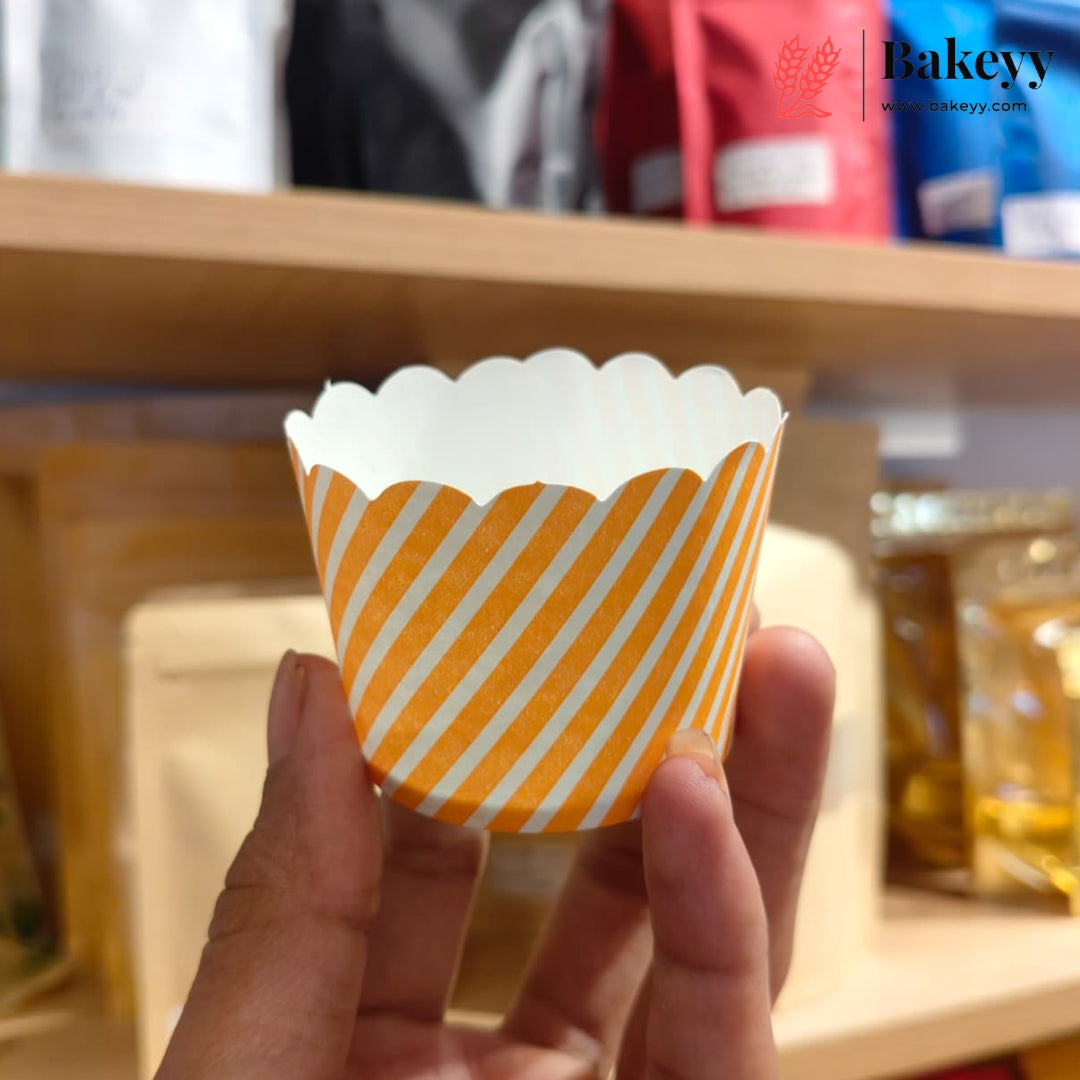 A hand holding a paper cup liner with orange and white diagonal stripes, with a bakery shelf blurred in the background. The Bakeyy logo is visible in the top right corner.