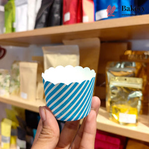A hand holding a blue and white striped paper cupcake liner in front of a blurred bakery shelf background with packaged goods.