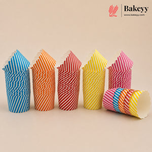 Six stacks of paper cupcake liners in assorted colors and stripe patterns, including blue and white, orange and white, red and white, yellow and white, pink and white, and a multicolor stack lying on its side. The Bakeyy brand logo is visible in the top right corner against a solid beige background.