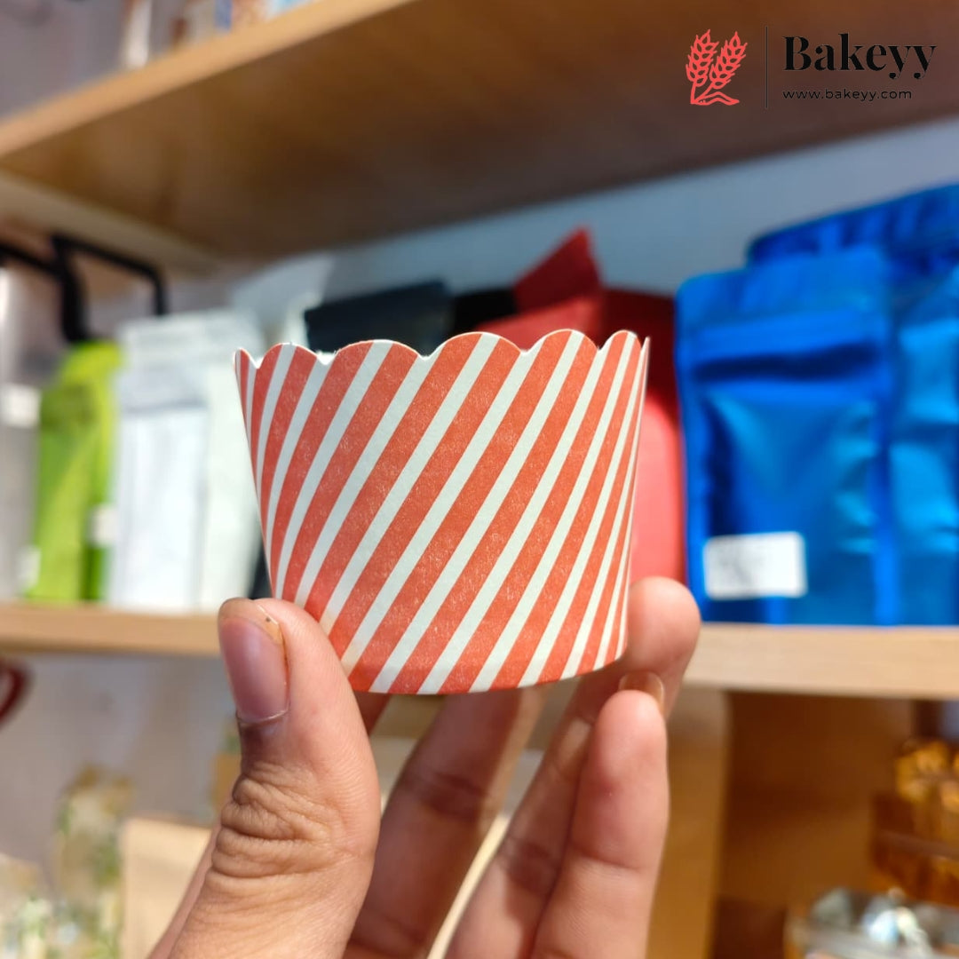 A hand holding a scalloped paper cupcake liner with diagonal orange and white stripes, set against a blurred bakery shelf background with a Bakeyy logo in the top right.