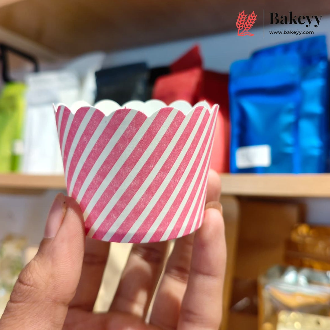A hand holding a pink and white striped scalloped paper cup liner, with a blurred bakery shelf background containing bags of coffee and other baking supplies. The Bakeyy logo is visible in the top right corner.