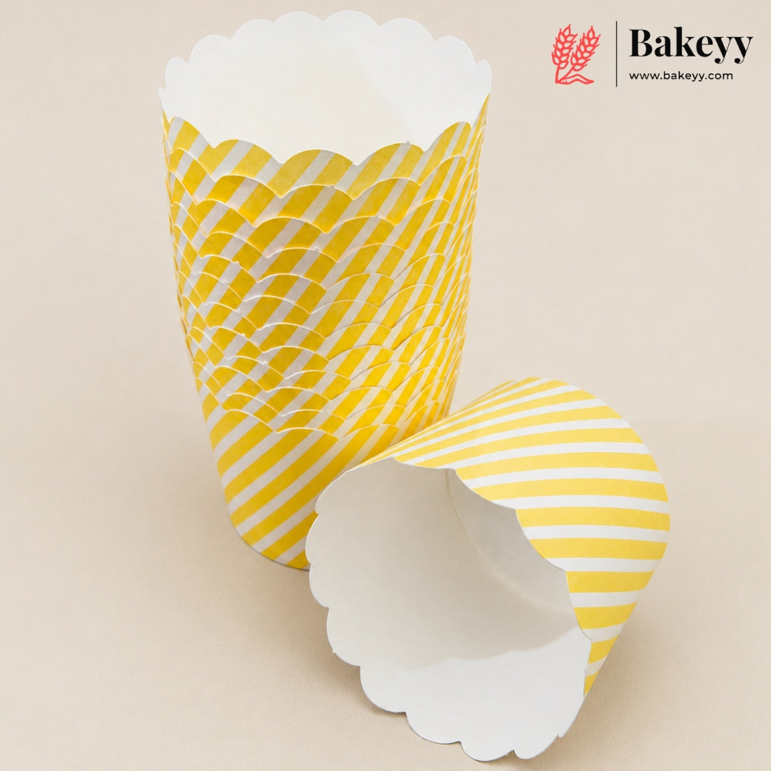 Three yellow and white patterned paper cupcake liners with scalloped edges, shown on a light beige background. One liner is standing upright, showing a yellow and white wavy pattern, while another is lying on its side showing a yellow and white diagonal stripe pattern.