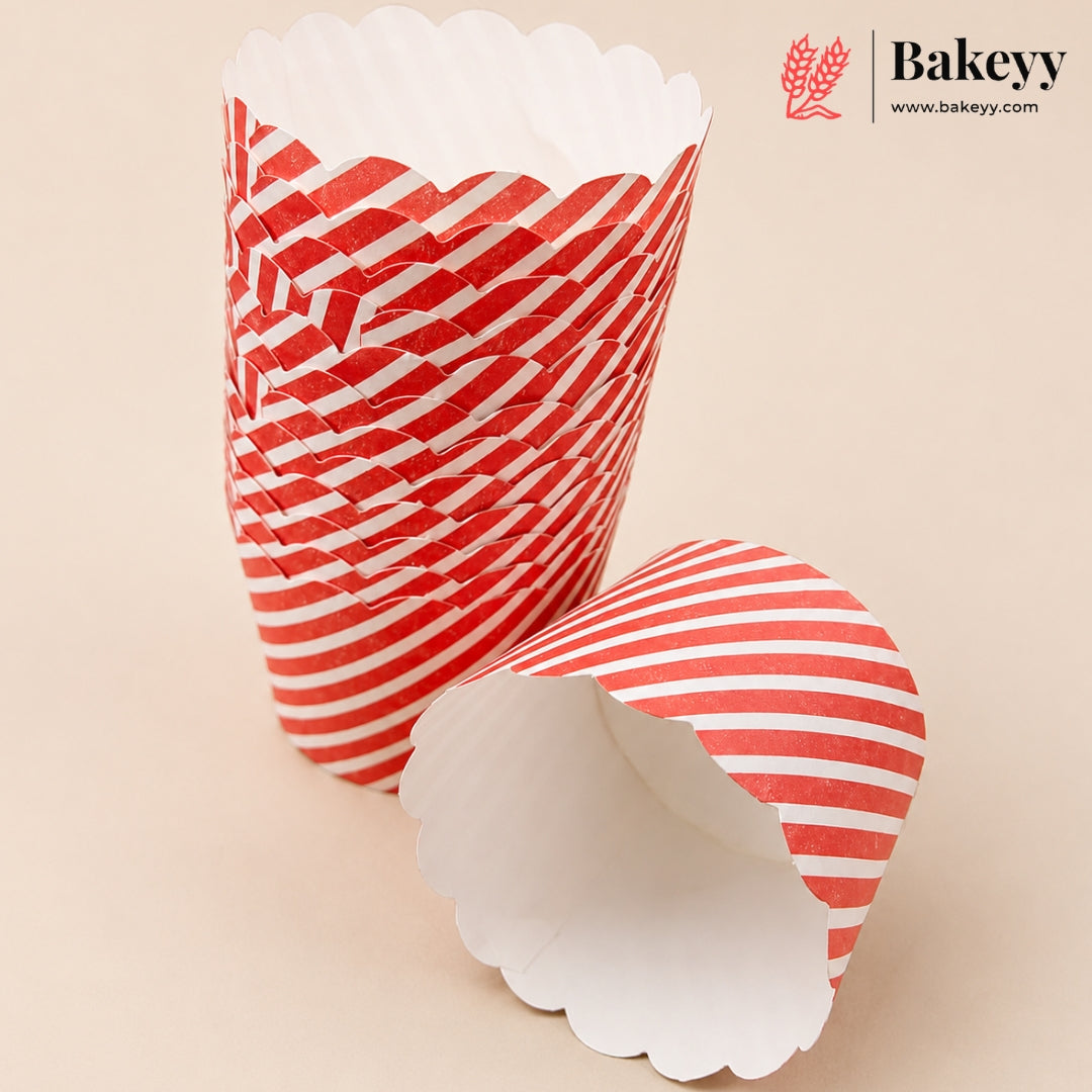 A stack of red and white striped paper cupcake liners with scalloped edges, with one liner lying on its side in front, on a light beige background. The Bakeyy logo is visible in the top right corner.
