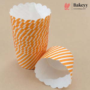 Two white paper cupcake liners with orange patterns, one standing upright and the other lying on its side, on a beige background. The Bakeyy logo is visible in the top right corner.