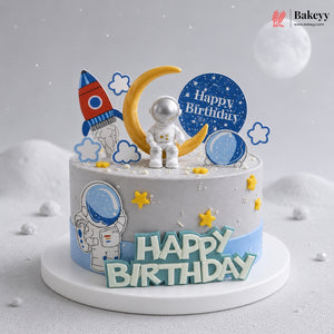 Space-themed birthday cake with astronaut, rocket, and moon decorations on a light gray background.