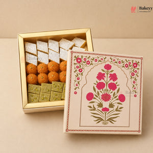 An open gold-toned gift box of Indian sweets and snacks displayed next to its decorated cream-colored lid, set against a solid beige background. The box contains white sugar-dusted sweets, orange chaklary, and green pistachio-filled sweets.