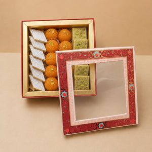 An open decorative gift box containing assorted sweets, including orange-colored round sweets, white rectangular sweets, and green pistachio-filled square sweets, with a red patterned lid leaning in front on a beige background.
