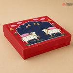 A square red gift box with a lid featuring a detailed illustration of two white cows with pink lotus flowers on a beige background.