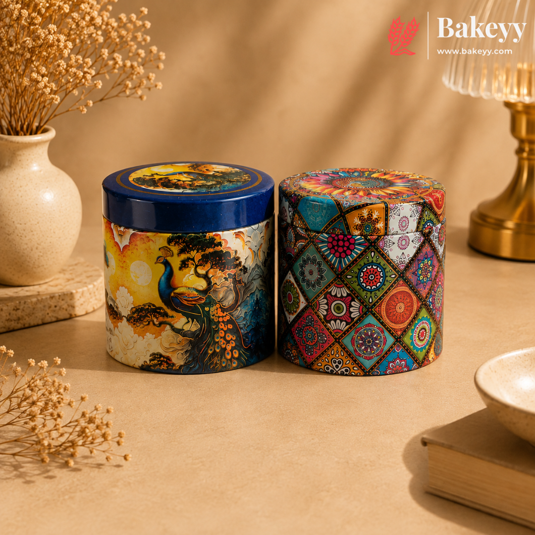 Two decorative cylindrical metal tins with lids, displayed on a beige tabletop with dried flowers, a vase, and a lamp in the background. The tin on the left has a blue lid and a peacock illustration on the body, while the tin on the right features a colorful geometric patchwork pattern.