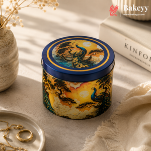 A decorative round metal tin with a blue lid and a colorful peacock landscape illustration, styled on a beige surface with a book, jewelry, and dried flowers in the background.