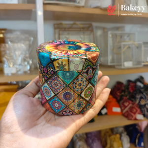 A hand holding a small cylindrical container decorated with a colorful, intricate geometric and floral pattern. The background shows a bakery or kitchen supply store with wooden shelves and glass jars.