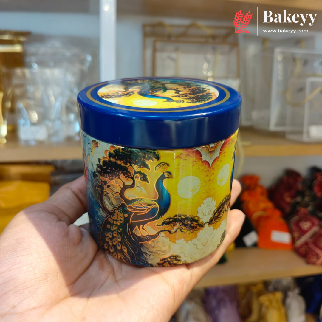 A hand holds a round decorative tin with a blue lid and a colorful peacock illustration on the side, set against a blurred store background with a Bakeyy logo in the top right corner.