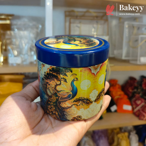 A hand holds a round decorative tin with a blue lid and a colorful peacock illustration on the side, set against a blurred store background with a Bakeyy logo in the top right corner.