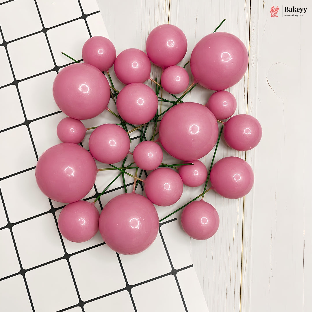 A cluster of glossy pink decorative beads or pom-poms on thin green stems, arranged on a white surface with a black grid pattern and a white wood plank background.