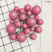A cluster of glossy pink decorative beads or pom-poms on thin green stems, arranged on a white surface with a black grid pattern and a white wood plank background.