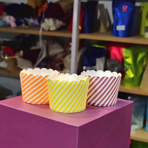 Three colorful striped cupcake liners on a pink surface with a blurred background of shelves.