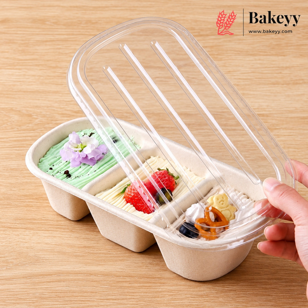 A three-compartment paperboard dessert tray filled with colorful cakes or pastries, shown on a wooden table with a hand lifting a clear plastic lid. The Bakeyy logo appears in the top right corner.